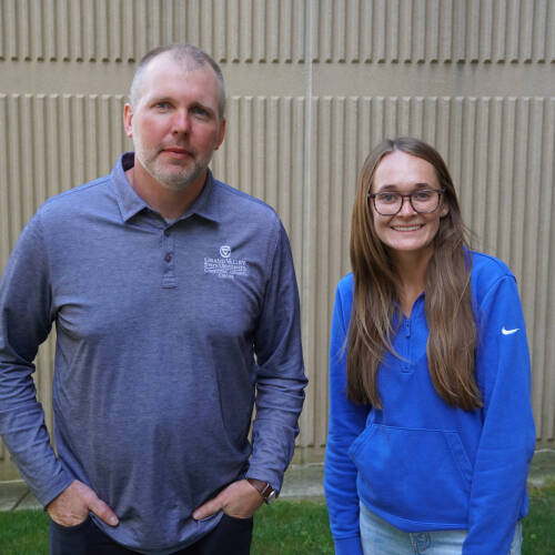 Colin DeKuiper and Emily Zeliasko, academic advisors in the GVSU College of Computing, standing outside against a tan textured wall. Colin is wearing a Grand Valley State University polo shirt, and Emily is wearing a blue pullover and glasses, smiling.